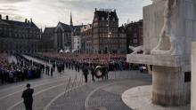 Official wreath-laying ceremony at the World War II monument on Dam Square, Amsterdam, the Netherlands. An orderly crowed is seated on a city square in front of brick buildings and a while statue and column representing the Dutch victims of World War II.