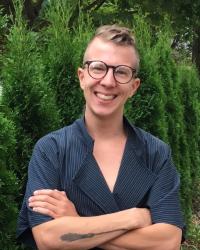 Image of young White male smiling and wearing glasses and a black tunic in front of green bushes.