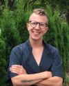 Image of young White male smiling and wearing glasses and a black tunic in front of green bushes.